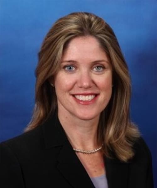 A woman with shoulder-length blonde hair wearing a black blazer, light purple blouse, and pearl necklace poses in front of a blue background.
