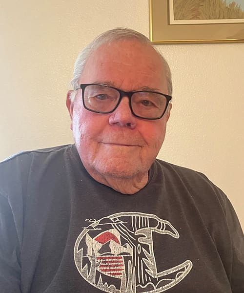 Older man with gray hair and glasses, wearing a black graphic t-shirt, sits indoors in front of a light-colored wall and framed artwork.